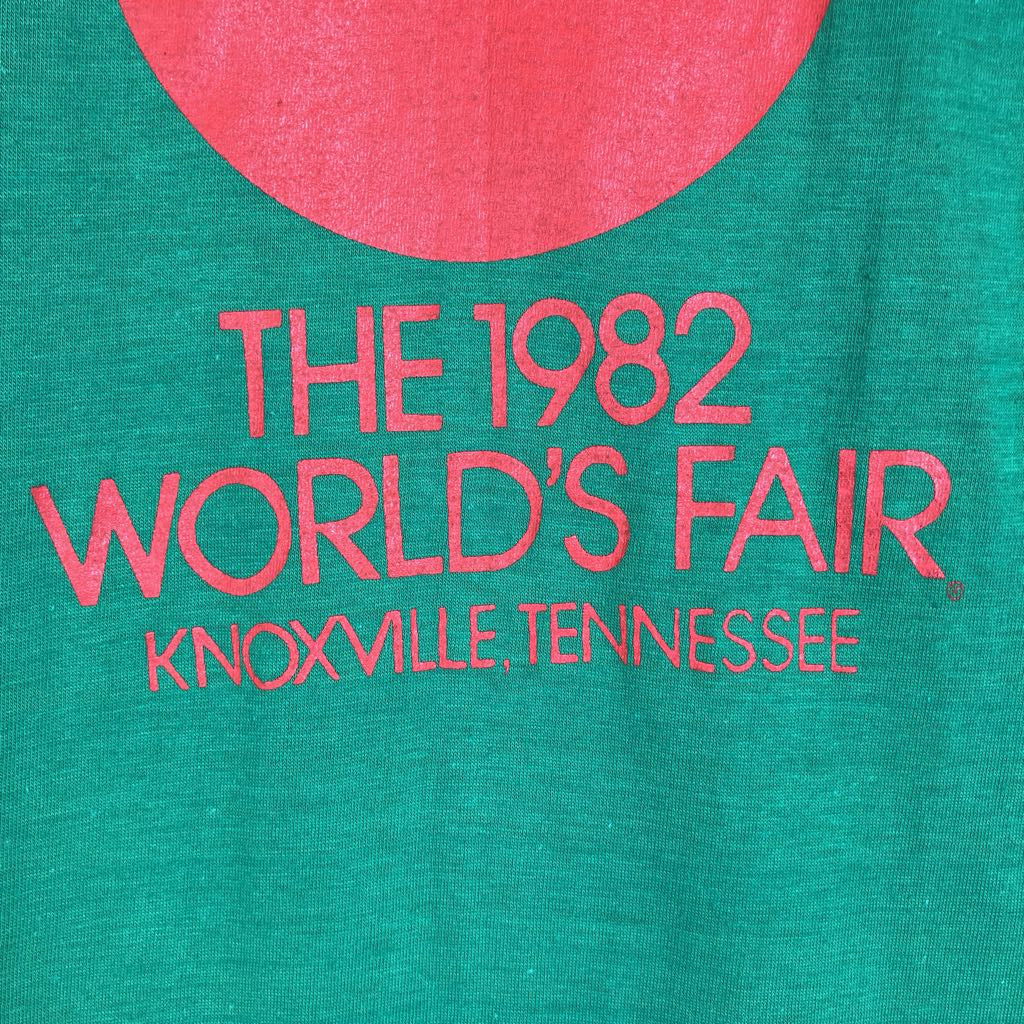 Close-up of a green fabric surface with a large pink circular graphic and pink text reading “THE 1982 WORLD’S FAIR KNOXVILLE TENNESSEE.”.