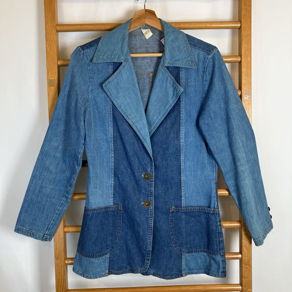A blue denim patchwork blazer jacket hanging on a wooden hanger from a wooden clothes rack against a plain white wall.