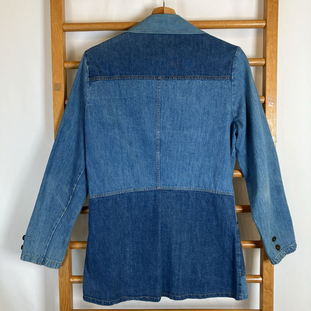 A blue denim jacket with a patchwork design, shown from the back hanging on a wooden hanger against a white wall with a wooden ladder behind it.
