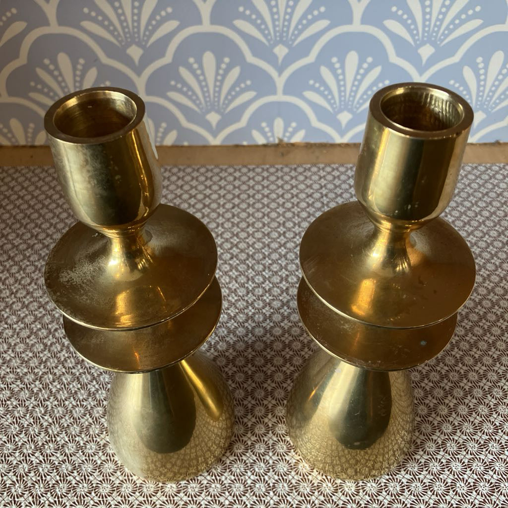 Two brass candle holders on a textured surface with a patterned wallpaper background.