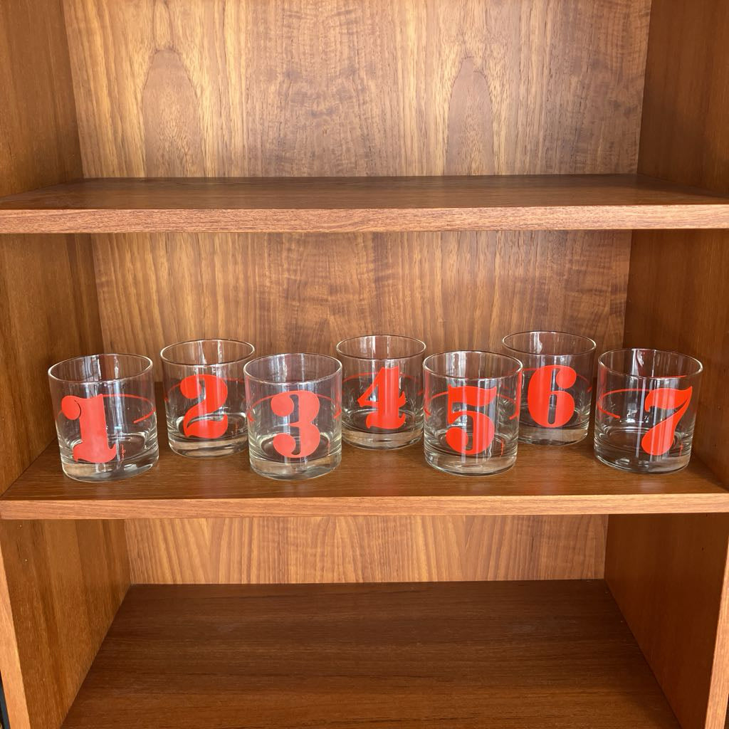 Set of 7 glass tumblers with red numbers on a wooden shelf.