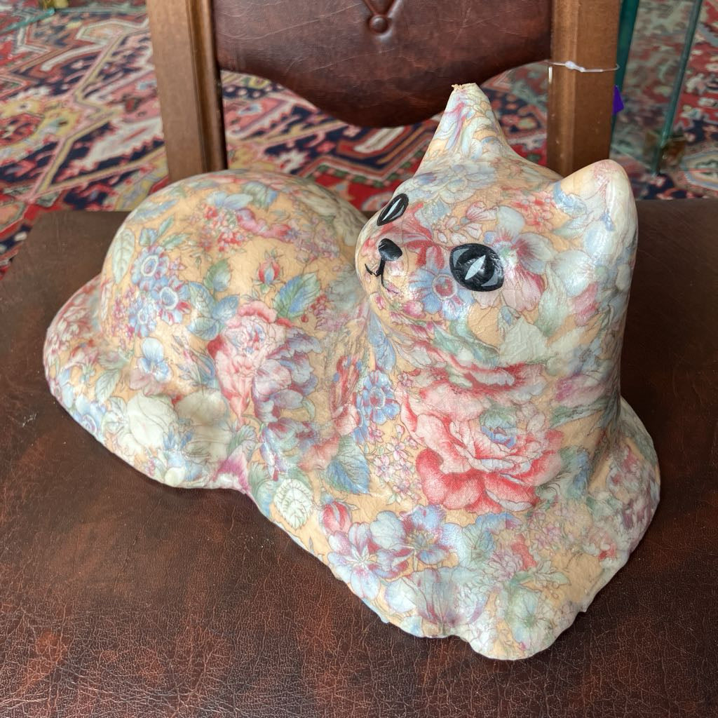 Floral-patterned cat-shaped object on a brown chair with a patterned rug in the background.