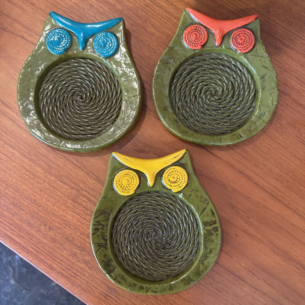 Three owl-shaped coasters with different colored eyes on a wooden surface.