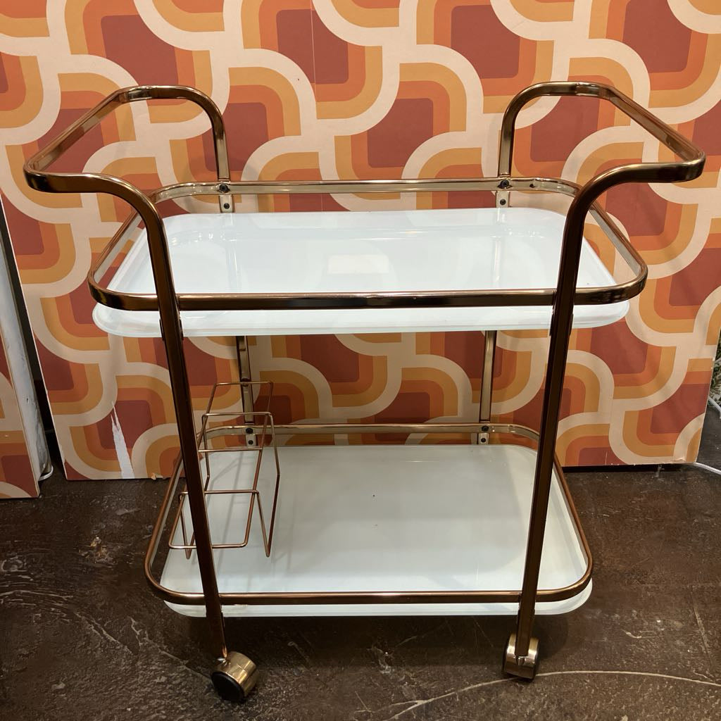 Vintage-style bar cart with gold frame and white shelves against a patterned wall.