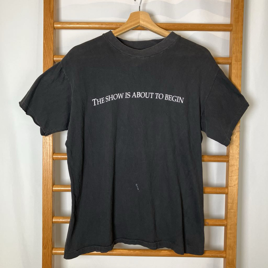 Black t-shirt with 'THE SHOW IS ABOUT TO BEGIN' text on a wooden hanger against a white wall.