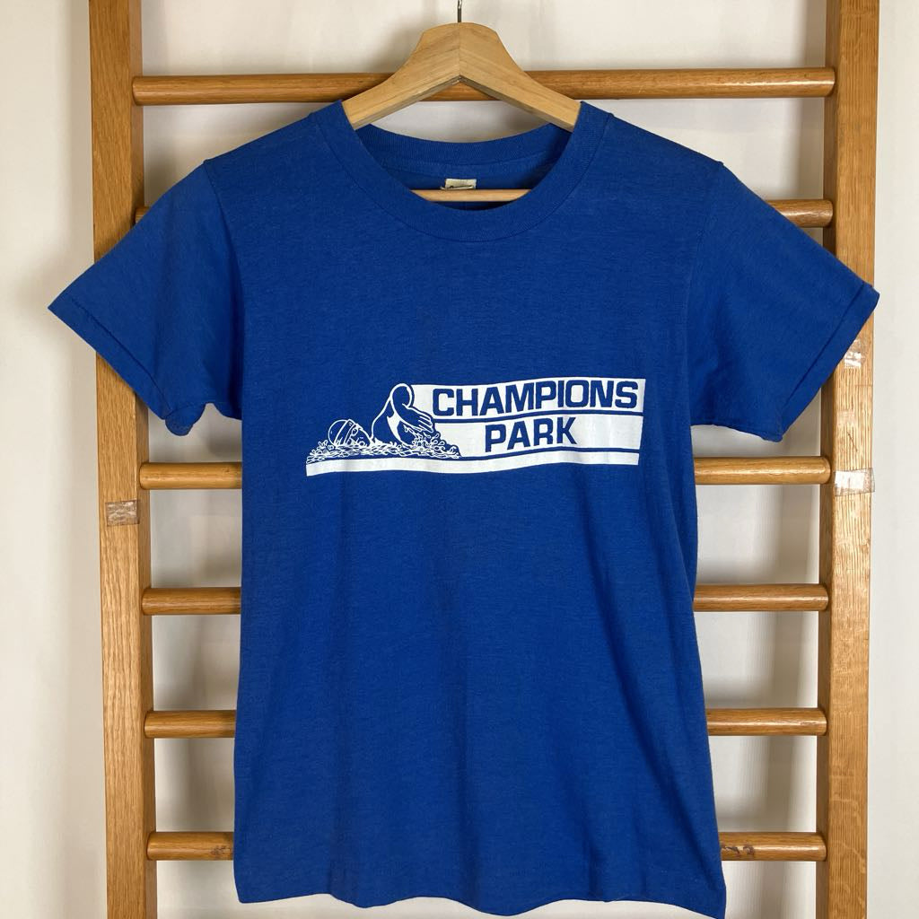 A royal blue short-sleeve t-shirt hanging on a wooden hanger against a wooden ladder. The shirt features a white graphic print on the chest that reads 'CHAMPIONS PARK' with a stylized skier.