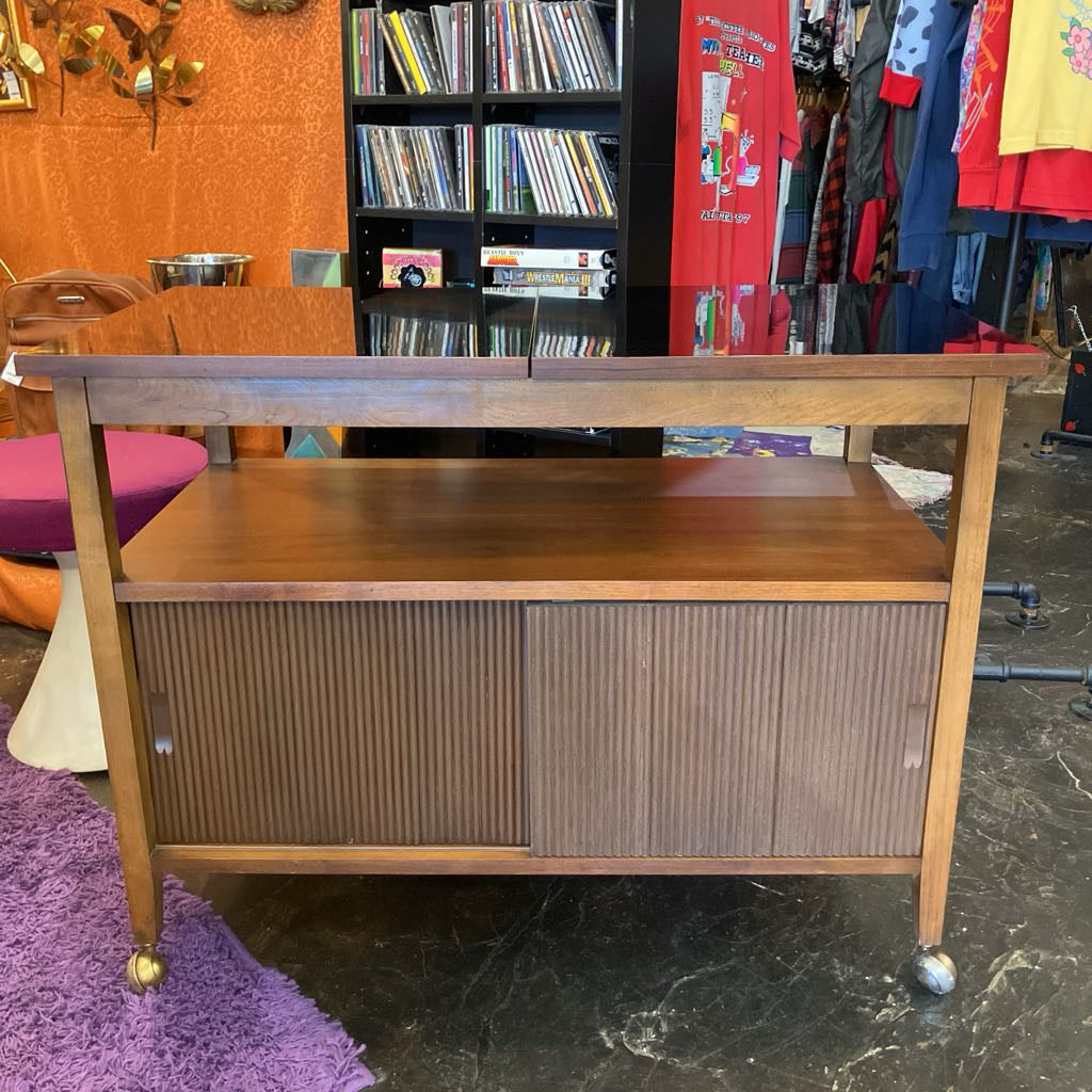 Wooden cabinet with sliding doors on wheels in a room with books and clothing.