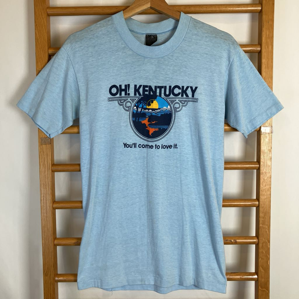 A light blue heather crewneck t-shirt hanging on a wooden hanger against a white wall, featuring a graphic print of the Ohio River Valley Museum in Louisville, Kentucky, with the text 'OH! KENTUCKY' and 'You'll come to love it.'.