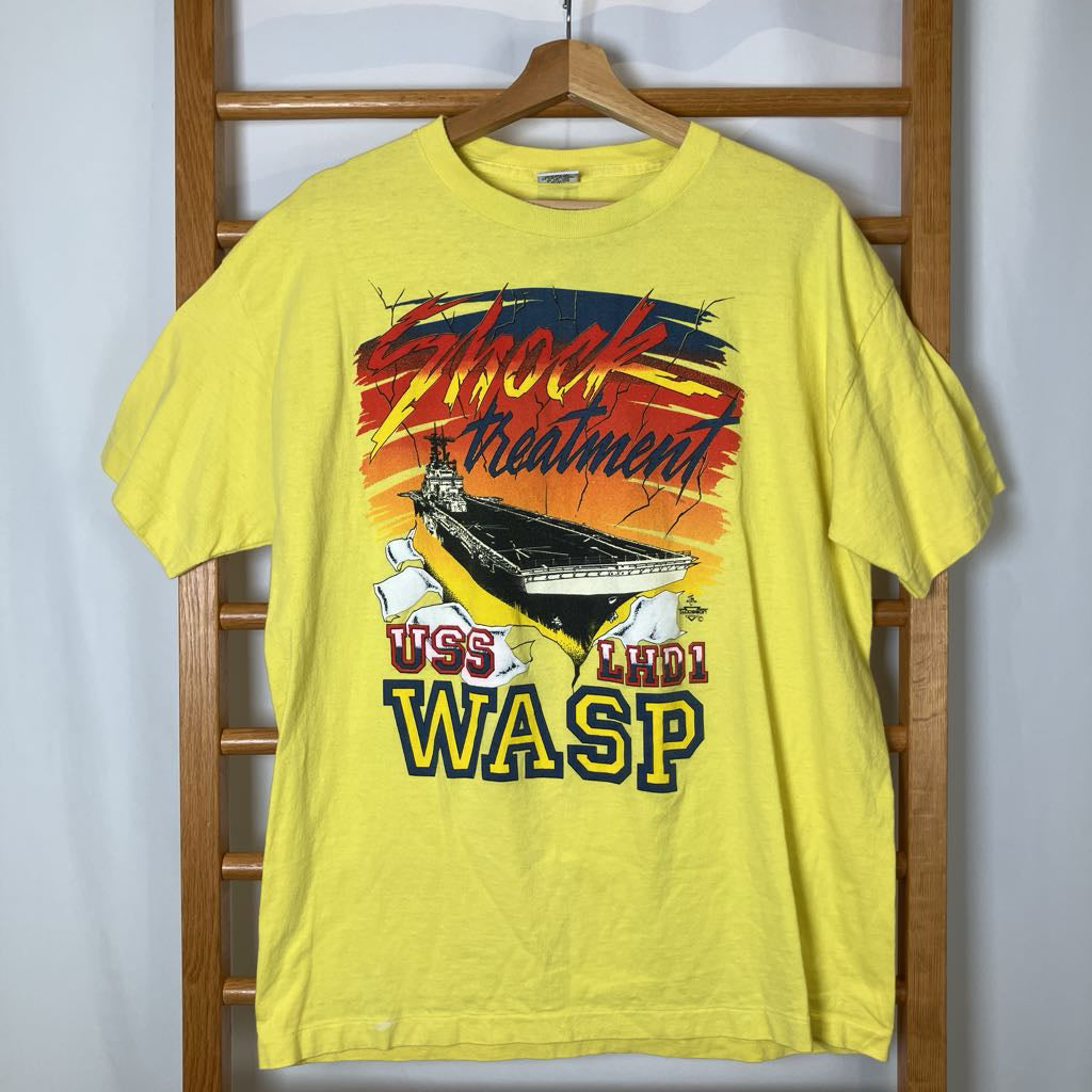 Yellow t-shirt with graphic design of USS LHD1 WASP on a wooden hanger against a white wall.
