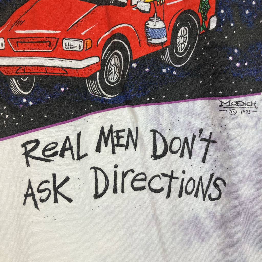 A close-up of a vintage graphic t-shirt featuring a red car on a starry black background above the text 'Real Men Don't Ask Directions' in black handwritten-style lettering on a white tie-dye fabric.