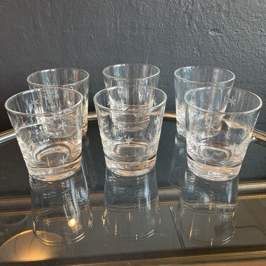 Six clear glass tumblers with a textured, etched pattern, arranged on a reflective dark tray against a dark gray textured wall.