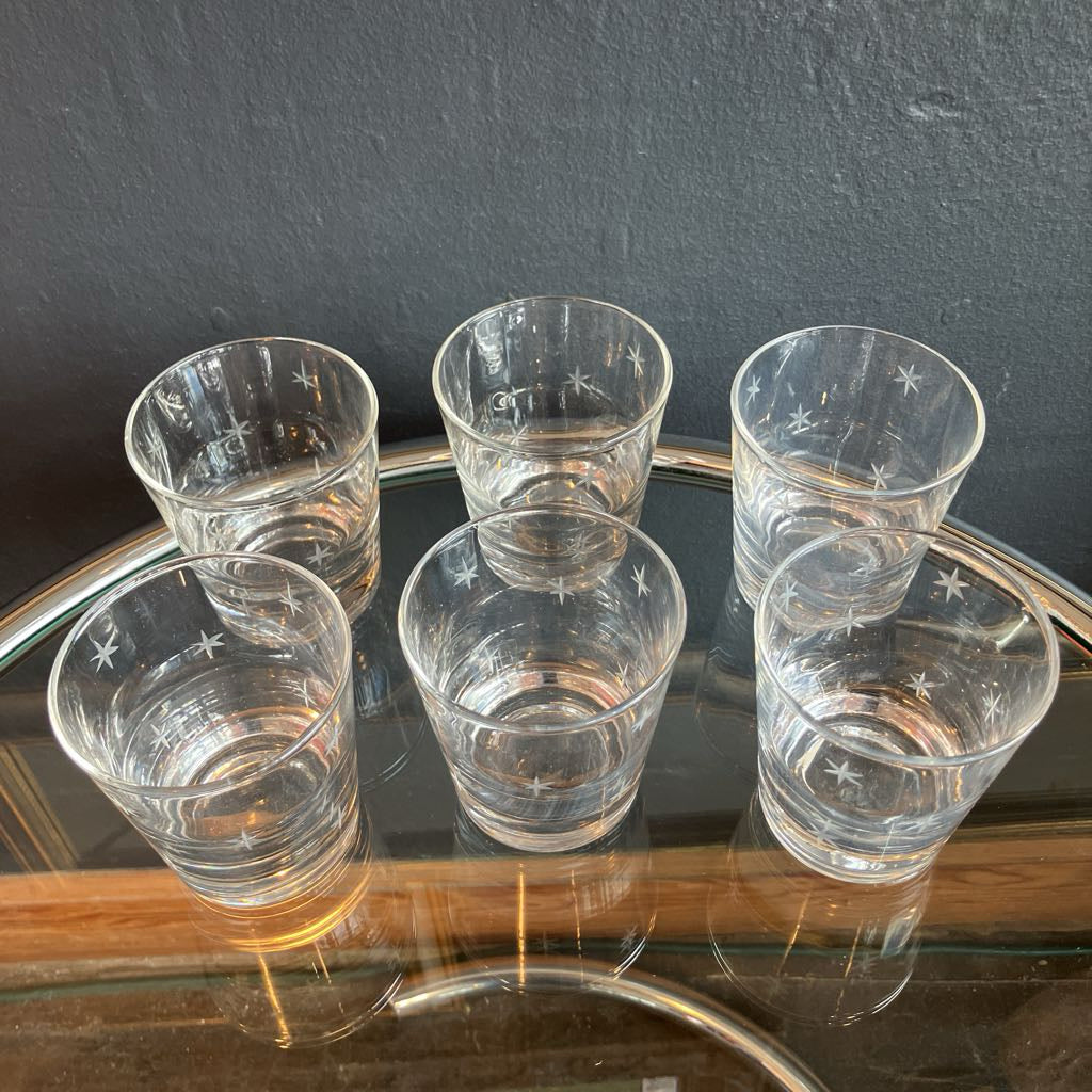 Six clear glass tumblers with etched star patterns, arranged on a glass tabletop against a dark gray wall.