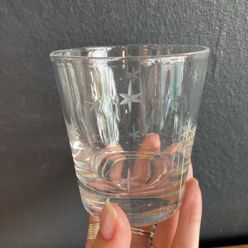A hand holding a clear glass tumbler with etched star patterns, set against a dark textured wall.