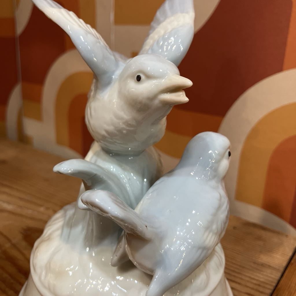 White ceramic birds on a decorative vase against a patterned wall.