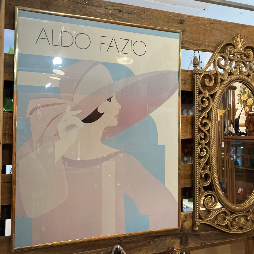 Framed poster of a woman with a large hat and 'Aldo Fazio' branding, next to an ornate mirror.
