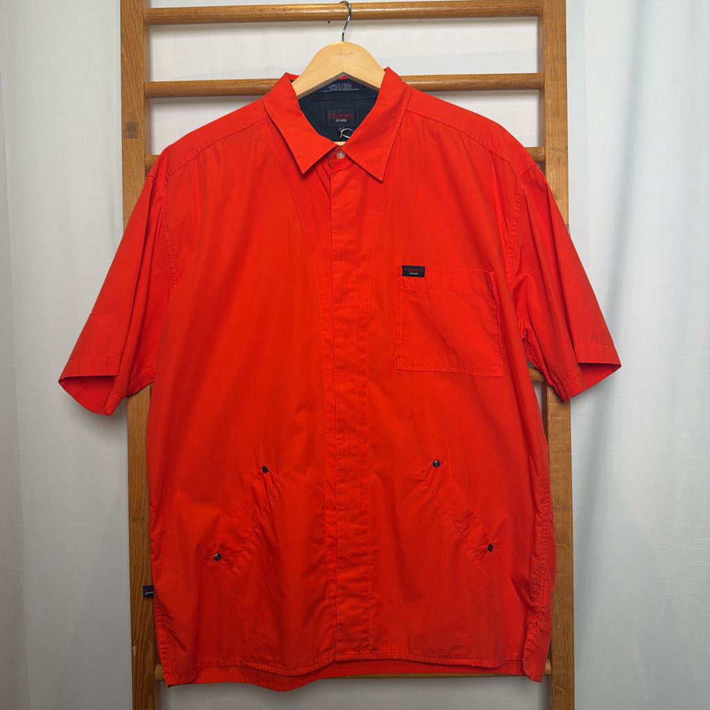 A bright red short-sleeve button-up shirt with a chest pocket and snap pockets, hanging on a wooden hanger on a wooden clothes rack against a white curtain background.