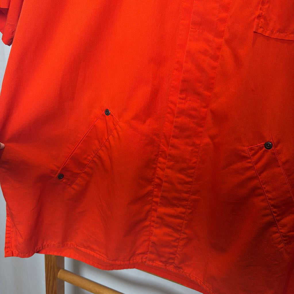 Close-up of a bright orange-red short-sleeve button-up shirt with two buttoned chest pockets and visible seam detailing, hanging against a light background.