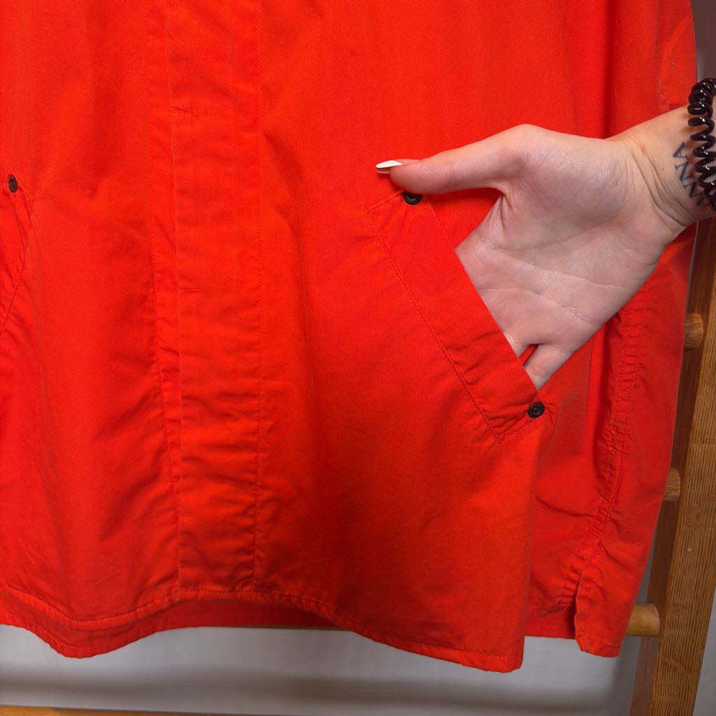 A close-up of a bright red-orange jacket or overshirt with a hand reaching into a side pocket, showing the fabric texture and snap-button details.