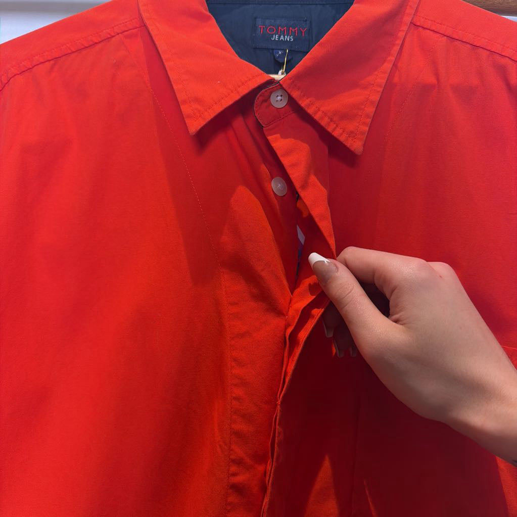 A close-up of a bright red Tommy Jeans button-up shirt being held open by a hand, showing the collar, buttons, and inner neck label.