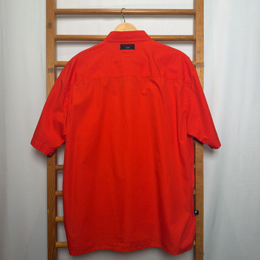 A bright red short-sleeve button-up shirt hanging on a wooden hanger against a white curtain backdrop, with a wooden ladder visible behind it.