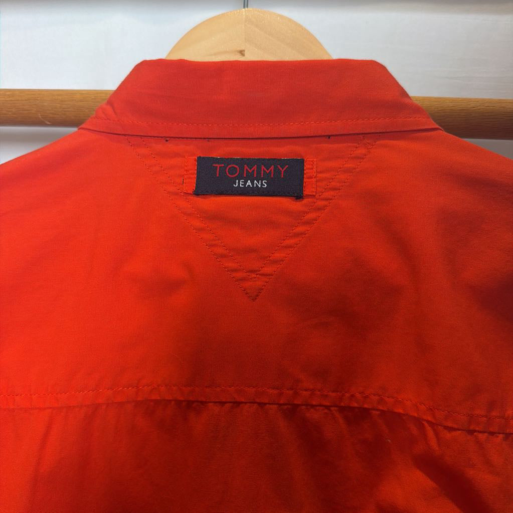 A close-up of a bright red-orange collared shirt jacket hanging on a wooden hanger, showing the Tommy Jeans brand label at the inner collar.