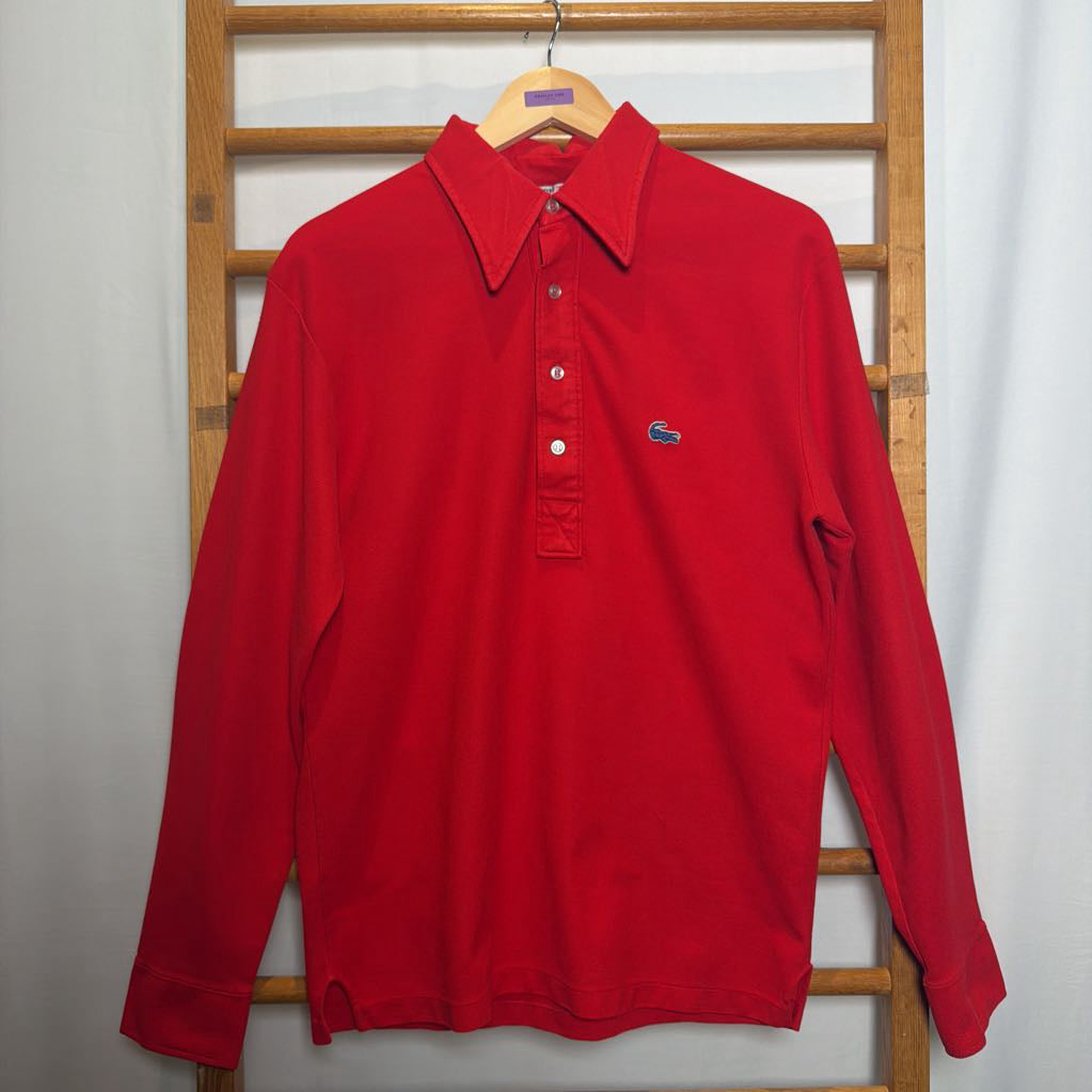 A red long-sleeve polo shirt with a three-button placket and the iconic crocodile logo on the left chest, hanging on a wooden hanger against a white curtain backdrop.