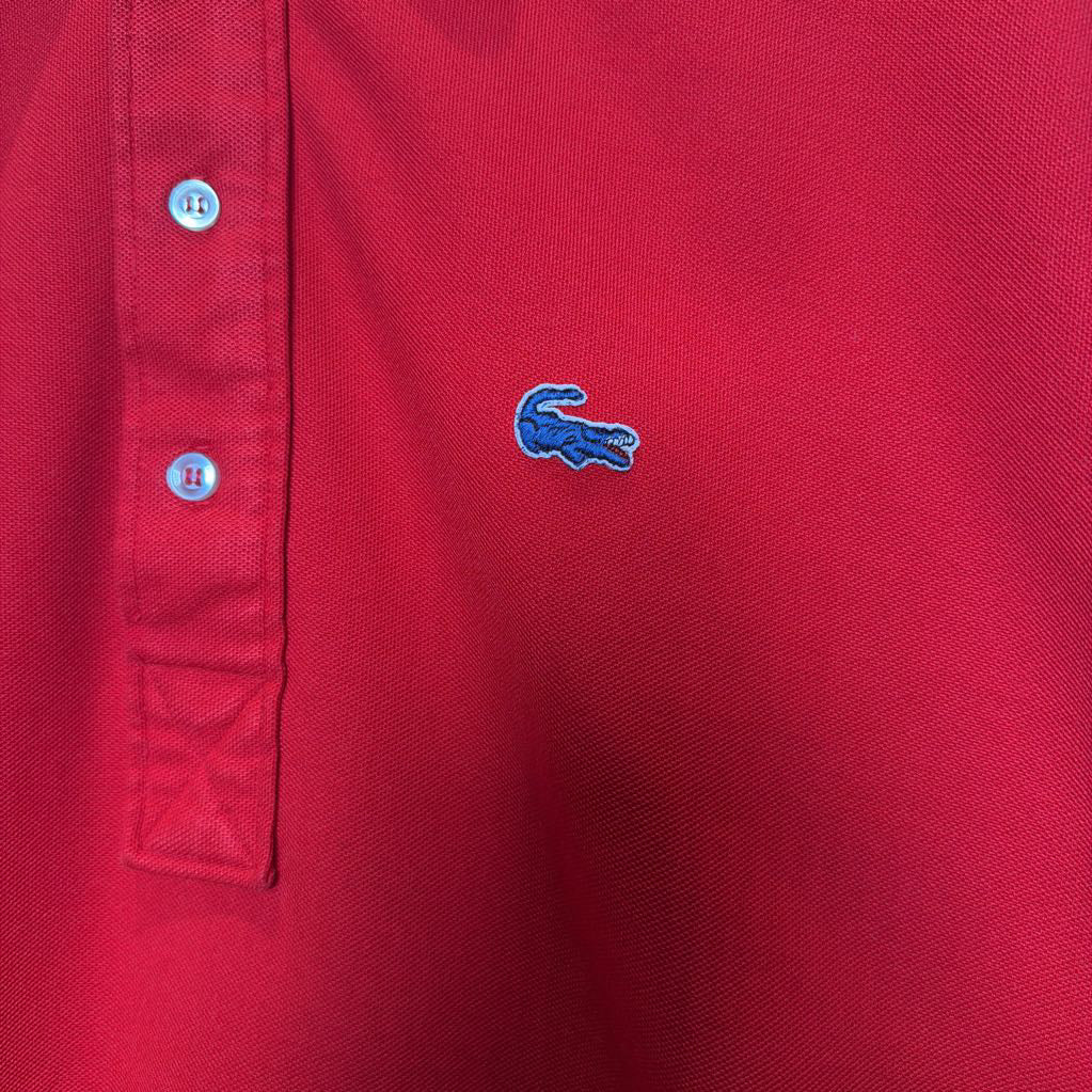 Close-up of a red polo shirt showing the three-button placket on the left side and the embroidered blue Lacoste crocodile logo on the chest.