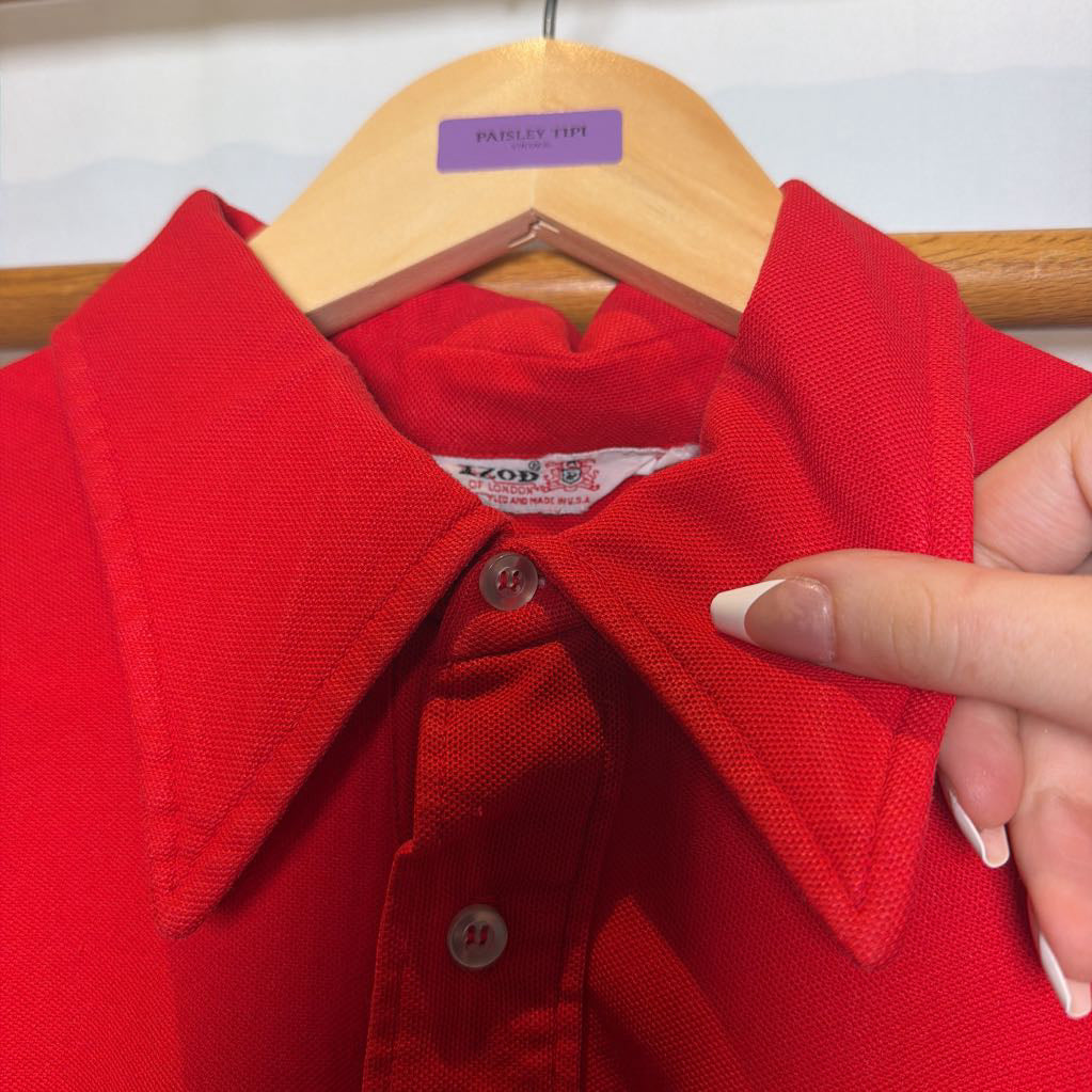 A close-up of a bright red polo shirt on a wooden hanger, with a hand holding the collar to show the fabric texture. The brand tag inside the collar reads 'AZOR'.