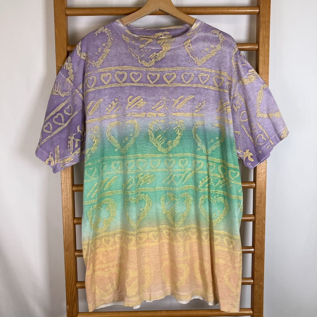 A short-sleeve t-shirt with a purple-to-green-to-yellow gradient and gold heart patterns, hanging on a wooden hanger against a white wall.