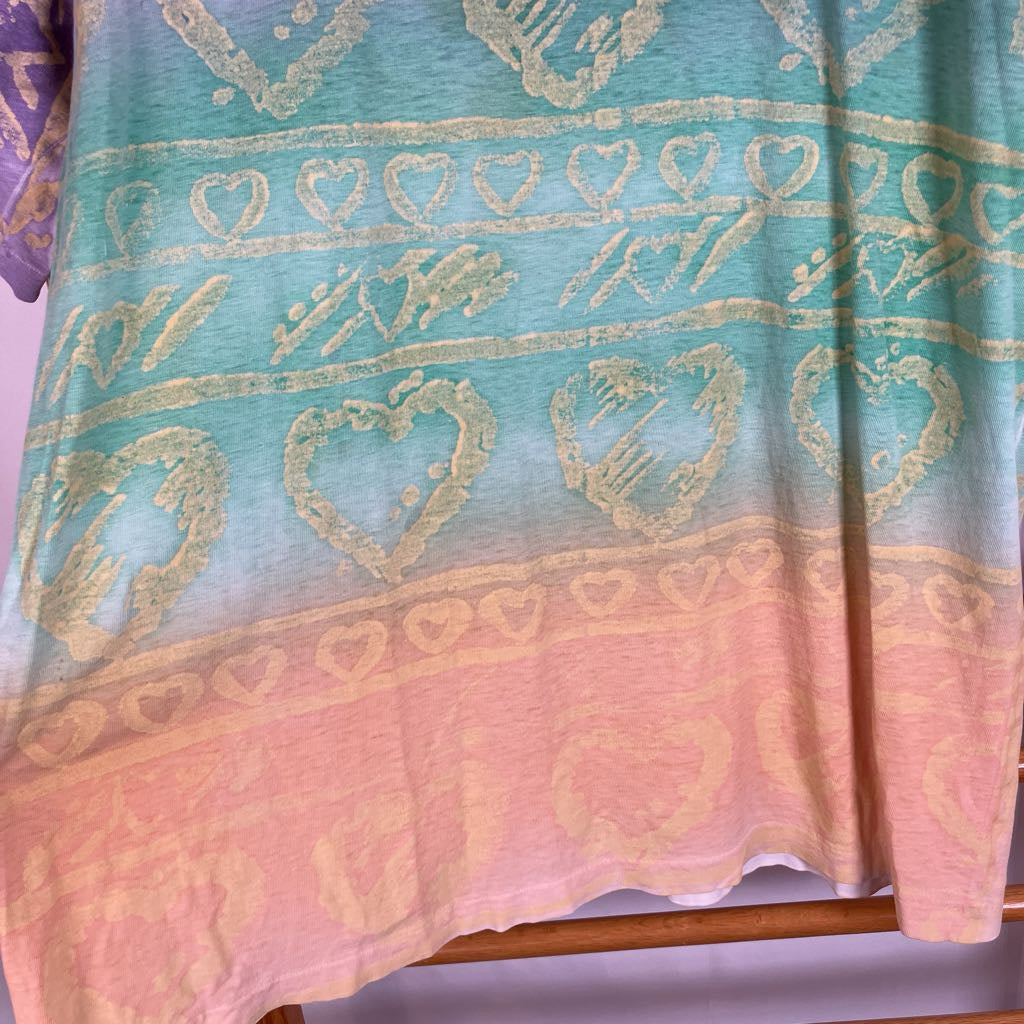 A close-up of a fabric garment or textile with a gradient design transitioning from light aqua blue at the top to soft pink at the bottom, decorated with distressed gold-toned heart and geometric patterns.