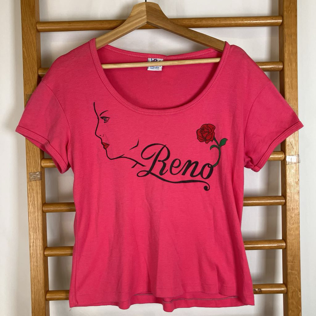 A bright pink scoop-neck t-shirt hanging on a wooden hanger against a wooden ladder. The shirt features a black graphic of a woman's profile, the name 'Reno' in cursive, and a red rose with green leaves.