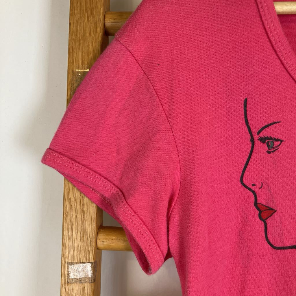A bright pink short-sleeve t-shirt hanging on a wooden hanger, featuring a minimalist black line art graphic of a woman's face on the right side.