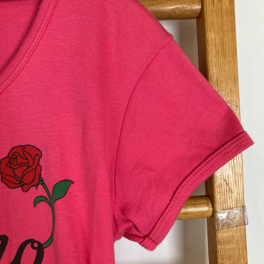 A close-up of a bright pink short-sleeve t-shirt featuring a red rose graphic with green leaves on the front, hanging against a wooden chair.