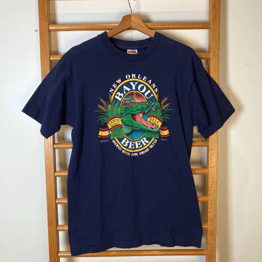 A navy blue short-sleeve graphic t-shirt hanging on a wooden hanger from a wooden ladder rack. The shirt features a colorful 'New Orleans Bayou Beer' alligator graphic on the chest.