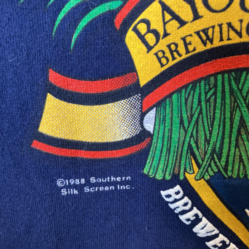 Close-up of a vintage graphic print on a dark navy blue fabric, featuring a yellow banner with 'BAYOU BREWING' in black text, green palm fronds, red and yellow stripes, and small white copyright text reading '©1988 Southern Silk Screen Inc.'.