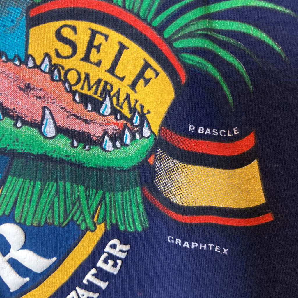 Close-up of a navy blue graphic print featuring a stylized crocodile head with green, yellow, red, and pink details, along with white text reading 'SELF OMNARY' and 'P. BASCLE'.