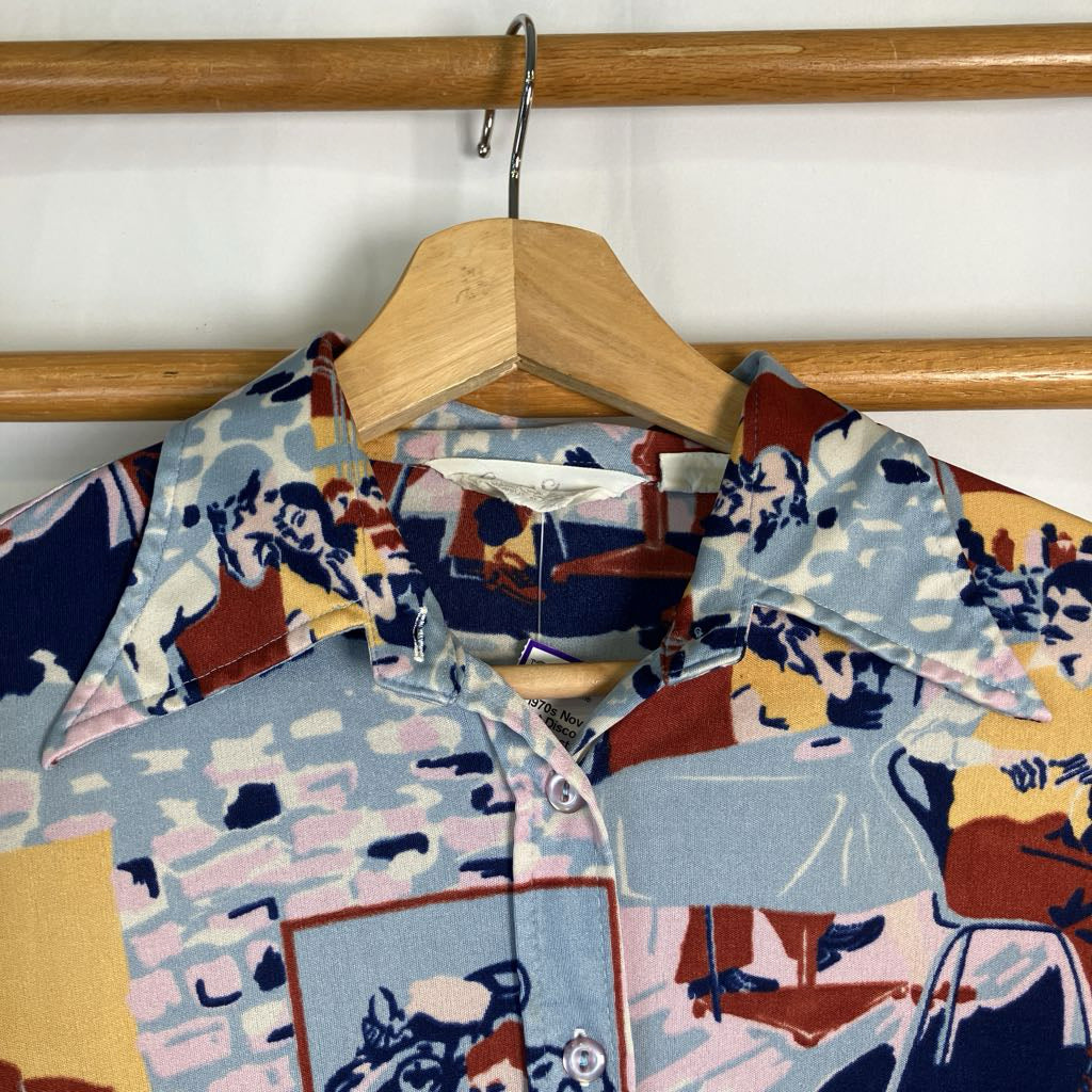 A short-sleeve button-up shirt with a busy collage-style print featuring figures, furniture, and abstract shapes in light blue, navy, red, yellow, and pink, hanging on a wooden hanger against a white wall.