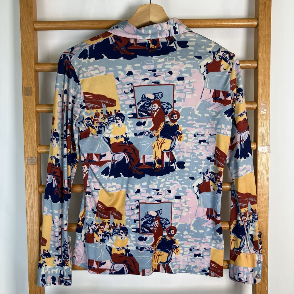 A long-sleeve collared shirt with a colorful collage print featuring cartoon-style figures, furniture, and abstract shapes, hanging on a wooden hanger against a white wall with a wooden ladder behind it.