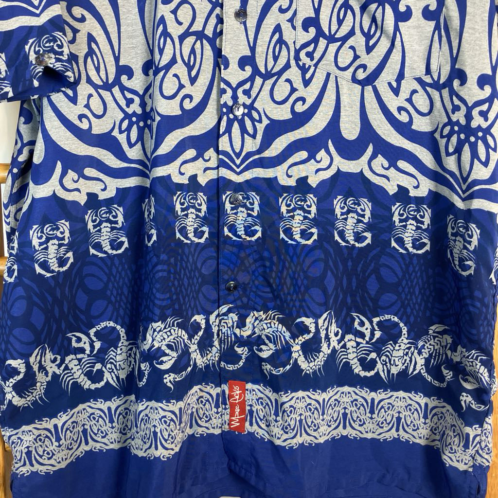 A close-up of a blue and white patterned button-up shirt or towel featuring traditional Chinese-style motifs including dragons, scorpions, and wave patterns, with a small red tag near the bottom.