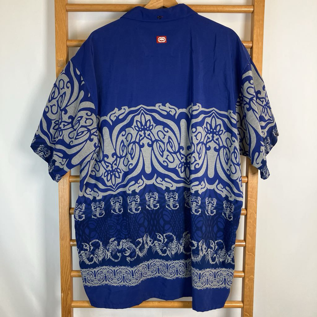 A royal blue short-sleeve button-up shirt with a large white and gray ornate batik-style pattern, hanging on a wooden hanger on a wooden chair against a white wall.