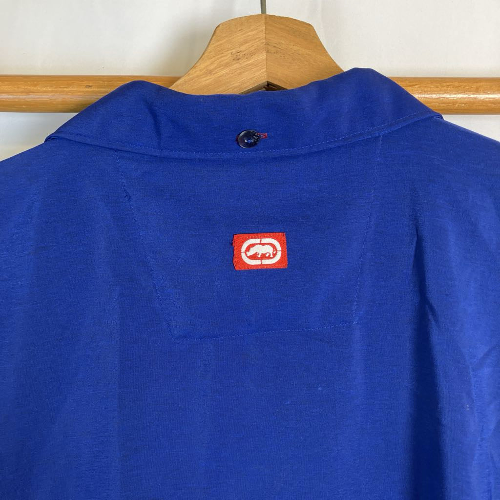 A royal blue knit top hanging on a wooden hanger, featuring a small red square patch with a white circular logo on the upper chest.