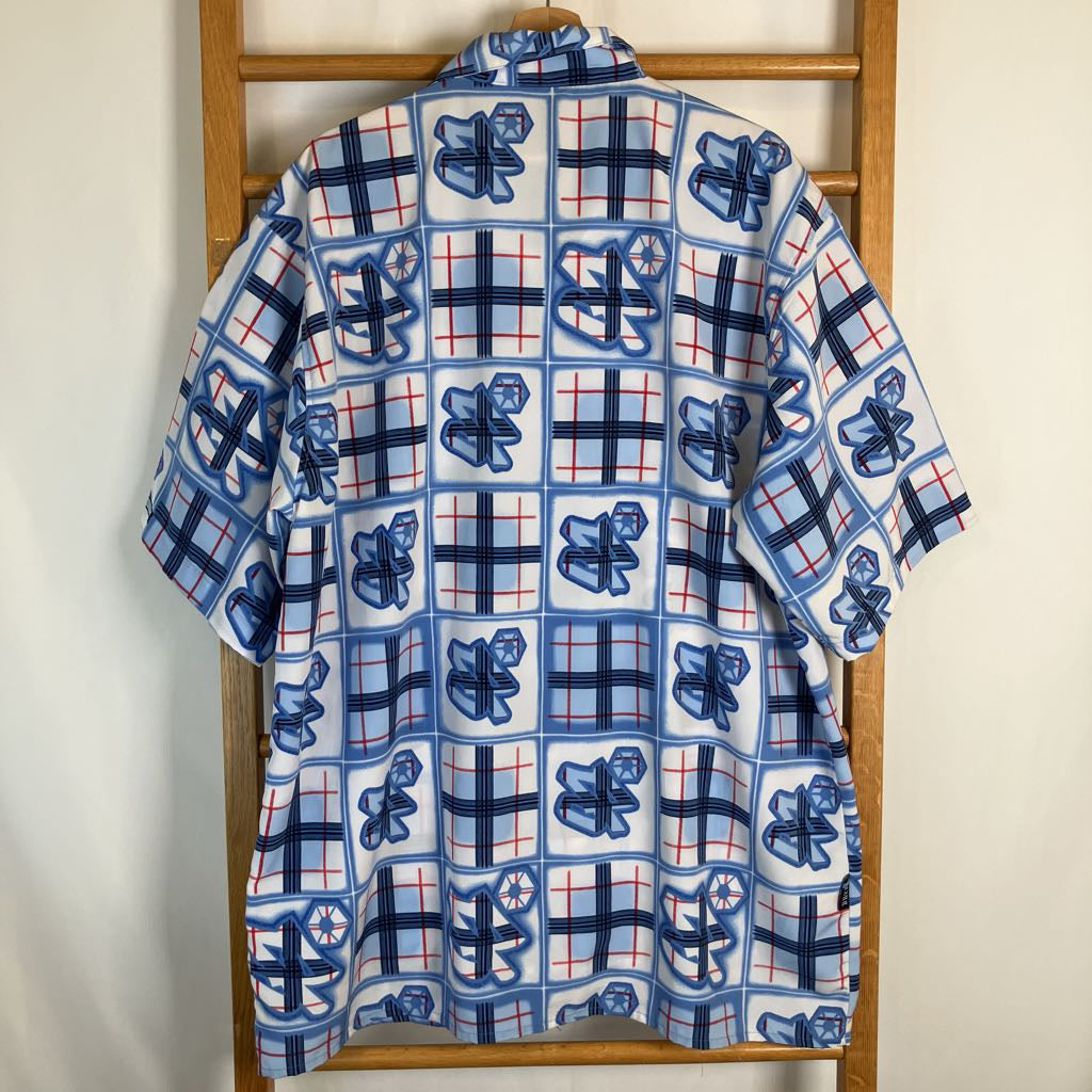 A short-sleeve button-up shirt with a blue, white, and red grid pattern featuring cartoon bear graphics, hanging on a wooden hanger against a plain off-white wall.