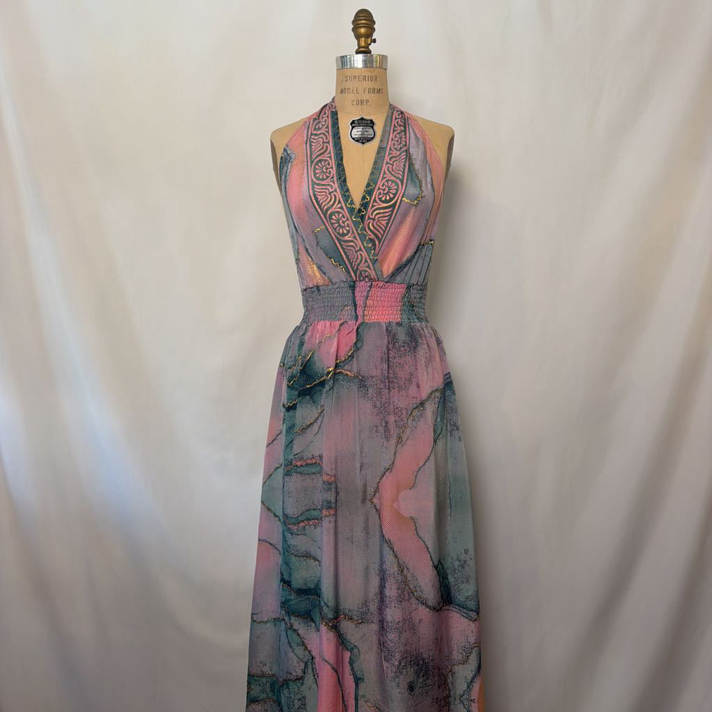 A sleeveless maxi dress with a V-neckline and waist tie, featuring a pink, teal, and gray abstract watercolor pattern, displayed on a dress form against a white fabric backdrop.
