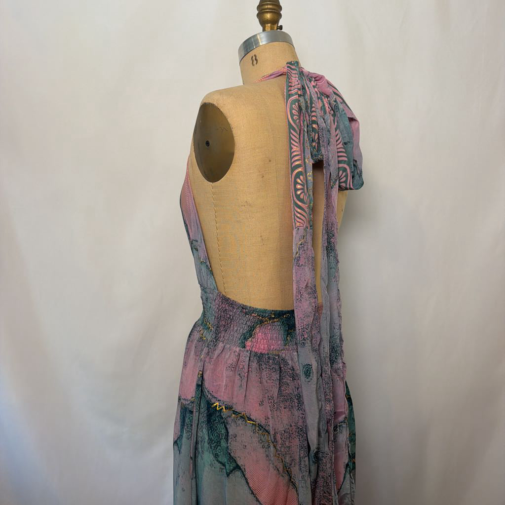 A sleeveless, backless dress displayed on a beige dress form mannequin against a white fabric backdrop. The dress features a pink, purple, and teal abstract pattern with a draped shawl or scarf detail over one shoulder.