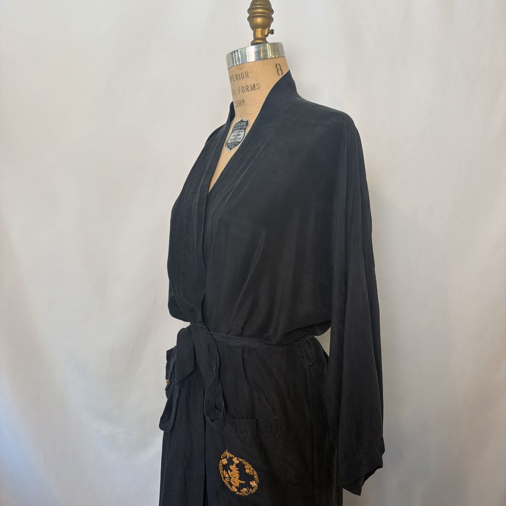 A black kimono-style robe displayed on a dress form mannequin against a light fabric backdrop. The robe has a self-tie belt and a circular gold-toned medallion patch near the lower hem.