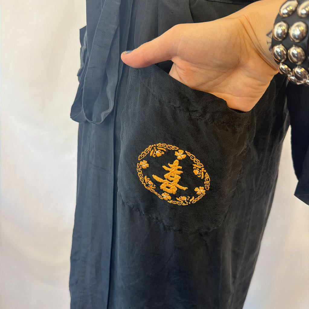 A close-up of a black fabric garment with a gold embroidered circular motif featuring a Chinese character, shown on a person wearing a silver beaded bracelet.