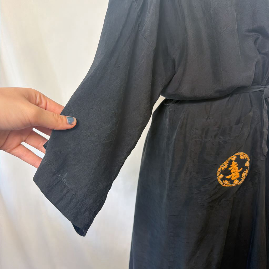 A close-up of a dark charcoal gray kimono-style robe with a hand holding up the sleeve to show the fabric texture and wide cuff. The robe features a gold embroidered circular motif on the side and is shown against a plain white fabric background.