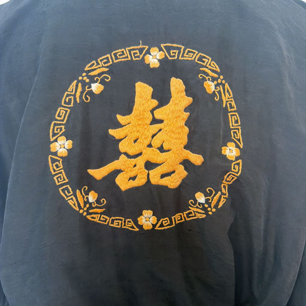 Close-up of a dark gray or black fabric garment featuring a large golden-yellow embroidered Chinese double happiness symbol inside a decorative circular border.