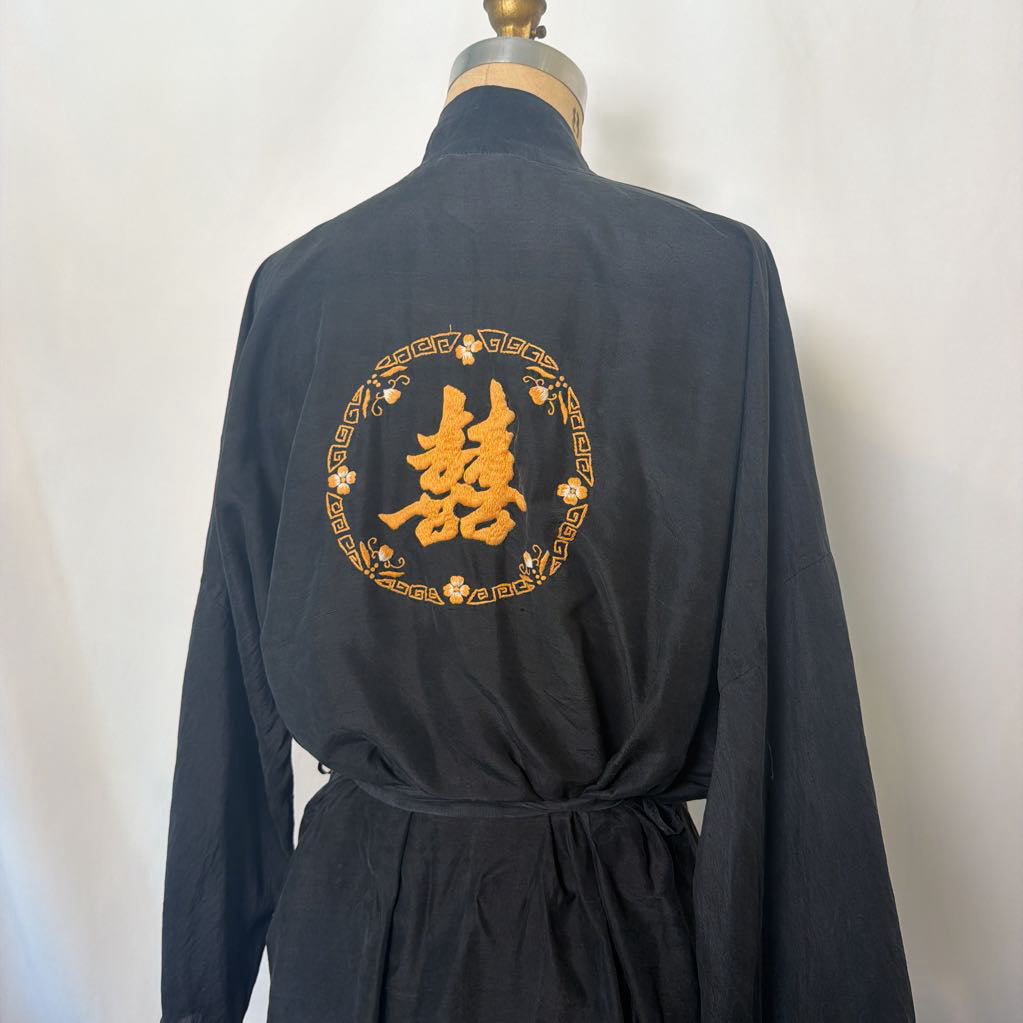 A black long-sleeve robe or kimono-style garment displayed on a dress form, showing the back with a large gold embroidered Chinese character and circular floral border motif.
