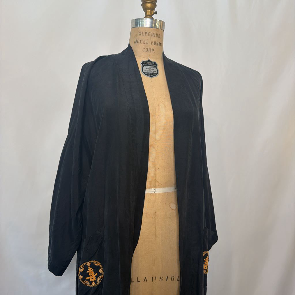 A black open-front kimono-style cardigan or robe displayed on a tan dress form mannequin against a light neutral backdrop. The garment has wide sleeves and gold embroidered circular motifs near the lower hem and sleeve cuffs.